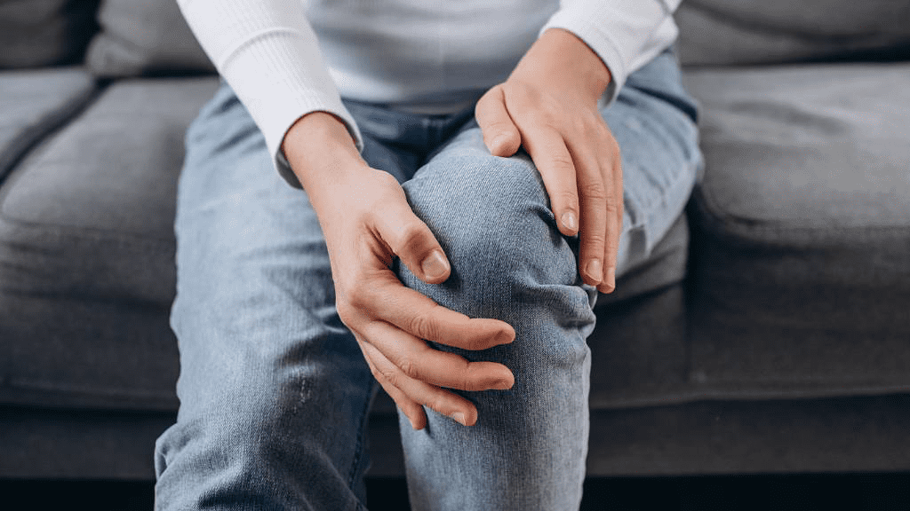 Person sitzt auf einem Sofa und hält sich mit beiden Händen das rechte Knie, offenbar wegen Schmerzen. Sie trägt eine hellblaue Jeans und ein weißes Langarmshirt. Symbolbild für Gelenkbeschwerden, Arthrose oder Knieprobleme.