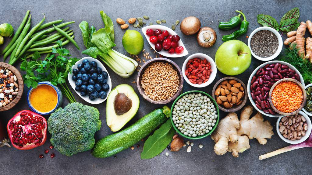 Eine Auswahl gesunder Lebensmittel, darunter frisches Gemüse, Obst, Hülsenfrüchte, Nüsse und Samen, auf einer dunklen Oberfläche arrangiert. Enthalten sind unter anderem Brokkoli, Blaubeeren, Avocado, Ingwer, Linsen, Mandeln und Kurkuma.