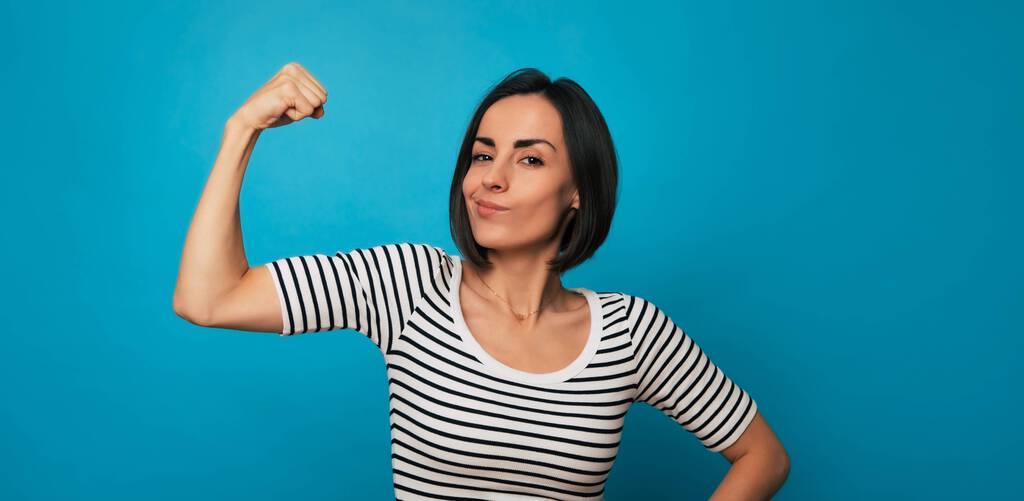 Eine junge Frau in einem gestreiften Oberteil beugt ihren Arm mit einem selbstbewussten Ausdruck vor einem blauen Hintergrund. Ihre Pose vermittelt Stärke, Kraft und Selbstvertrauen.