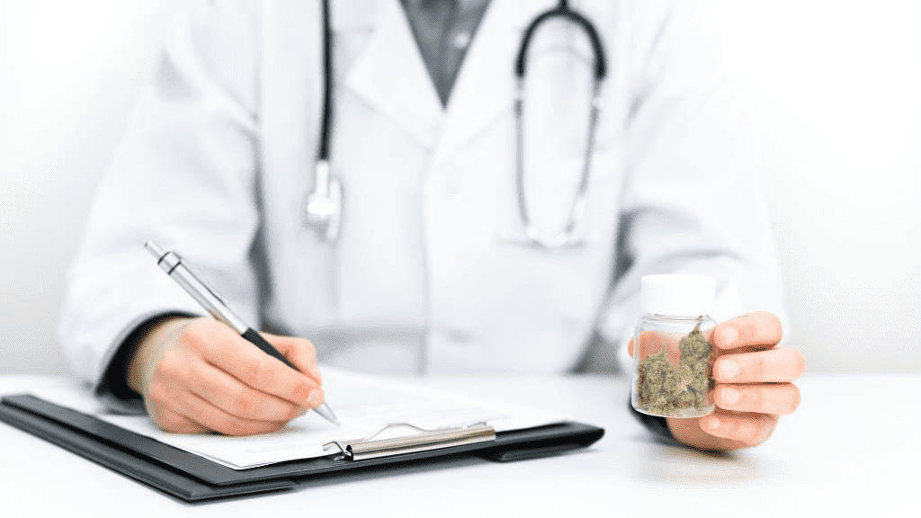 Arzt in weißem Kittel mit Stethoskop hält Glasbehälter mit medizinischem Cannabis und macht sich Notizen – Symbolbild für Cannabis auf Rezept.