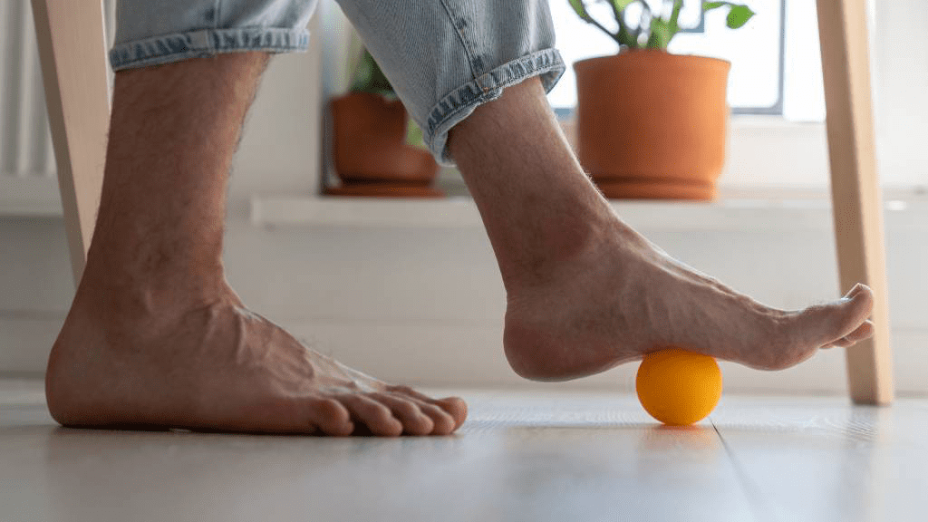 Eine Person führt eine Fußmassage mit einem gelben Ball durch, während sie barfuß auf dem Boden steht. Die Übung dient der Fußgesundheit und kann helfen, Fehlstellungen vorzubeugen und Verspannungen zu lösen.