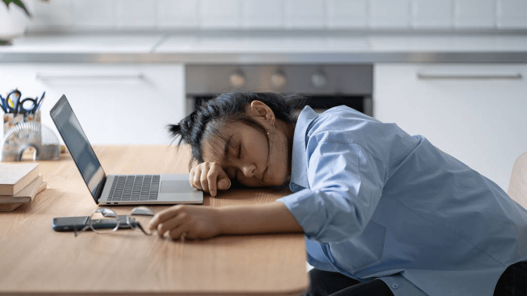 Frau mit braunen Haaren und blauem Hemt schläft. Ihr Kopf liegt auf auf der Tischplatte. Vor ihr steht ein Laptop.