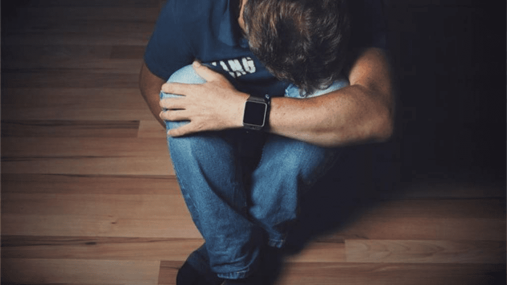 Ein Mann sitzt zusammengekauert auf dem Boden im Halbdunkeln und legt seine Stirn auf seinem Knie ab.