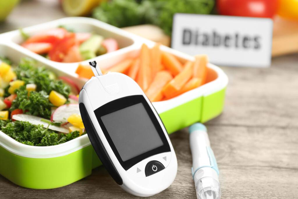 Blutdruckmessgerät, Insulinspritze und grüne Brotdose mit drei Fächern, darin Salat, kleingeschnittene Möhren und weiteres Gemüse. Im Hintergrund ein Schild mit Aufschrift "Diabetes".
