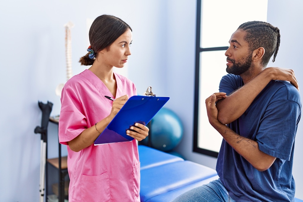 Eine gute Behandlung erfolgt immer auf Augenhöhe. | | © Krakenimages - stock.adobe.com Eine Chiropraktikerin hört ihrem Patienten aufmerksam zu. Sie hält ein Klemmbrett für Notizen in der Hand und macht sich Notizen.