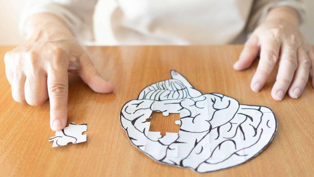 Eine ältere Frau sitzt am Tisch und berührt ein Puzzleteil, dass aus einem Papierbild eines Gehirns ausgeschnitten wurde. Sinnbild für Demenz.