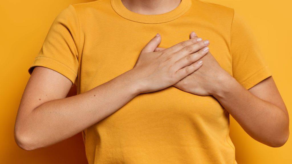 Eine Frau fasst mit beiden Händen an ihr Herz. Sie trägt ein gelbes T-Shirt und steht vor einem gelben Hintergrund.