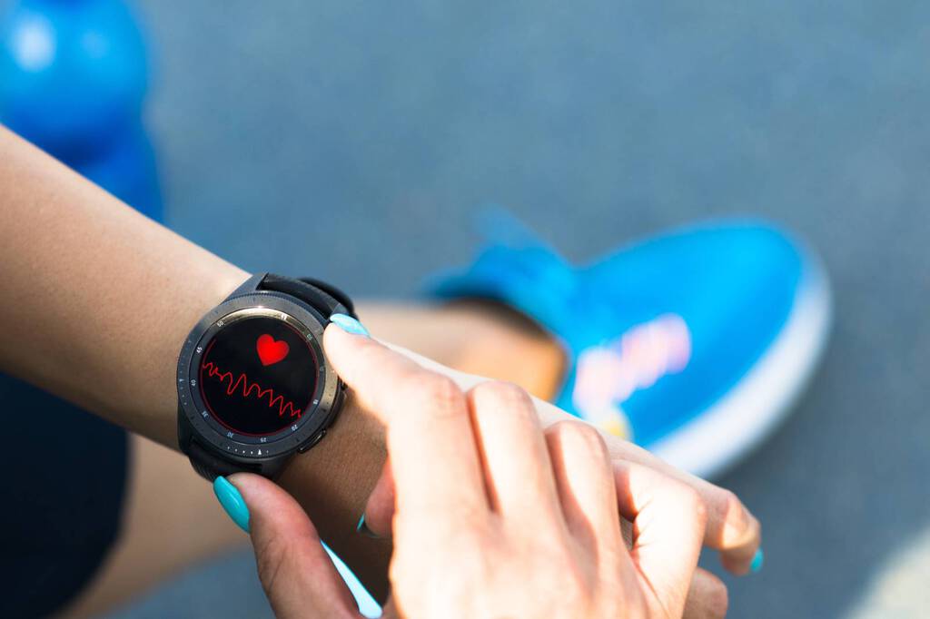 Smartwatch an einem Arm zeigt Herz und Herzfrequenz. Darunter sieht man blaue Turnschuhe und eine blaue Wasserflasche auf Asphalt.