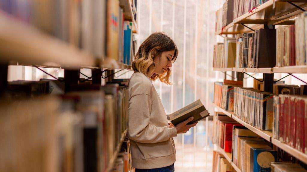 Eine Frau liest ein Buch. Sie steht zwischen zwei Buchreihen in einer Bibliothek.