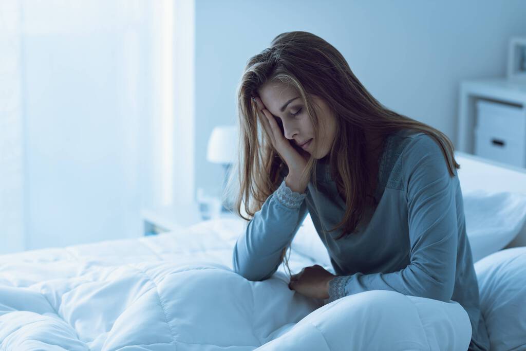 Eine erschöpfte Frau sitzt aufrecht im Bett. Sie stützt ihren Kopf mit ihrer rechten Hand ab.