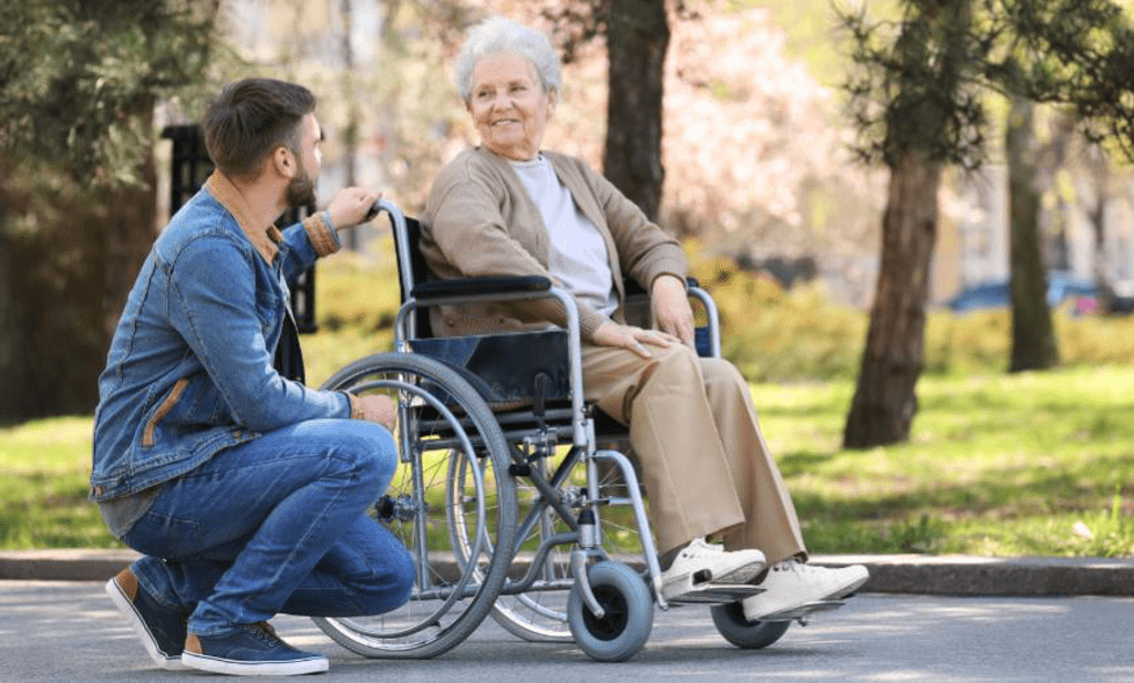 Eine lächelnde Seniorin im Rollstuhl in einem Park. Neben ihr kniet ein junger Mann und sieht ihr in die Augen.