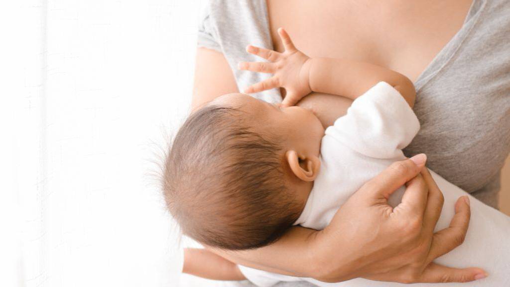 Eine Frau, stillt ein Baby an ihrer Brust. Das Kind liegt in ihrem Arm, trägt ein weißes Oberteil und saugt an der Brust. Die Szene wirkt nah, ruhig und innig.