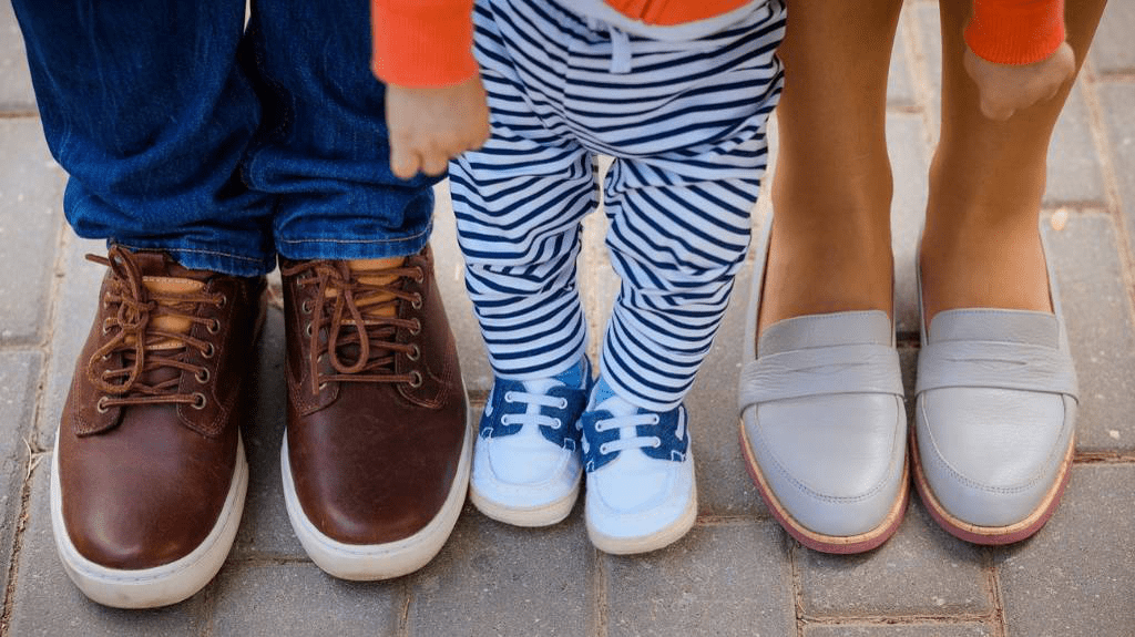 Drei Paar Füße in Schuhen. Links: Lederschnürschuhe mit Jeansbeinen. Mitte: Kleinkind in blau-weißen Sneackern mit blau-weiß geringelter Hose. Rechts: gräuliche Halbschuhe und nackte Knöchel.