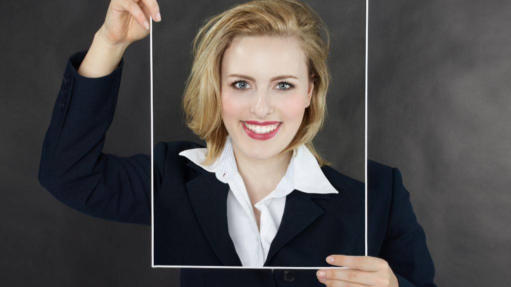 Eine Frau im Businesskostüm. Ihren Kopf verdeckt ein Bild Ihres lächelnden und fröhlichen Gesichts.