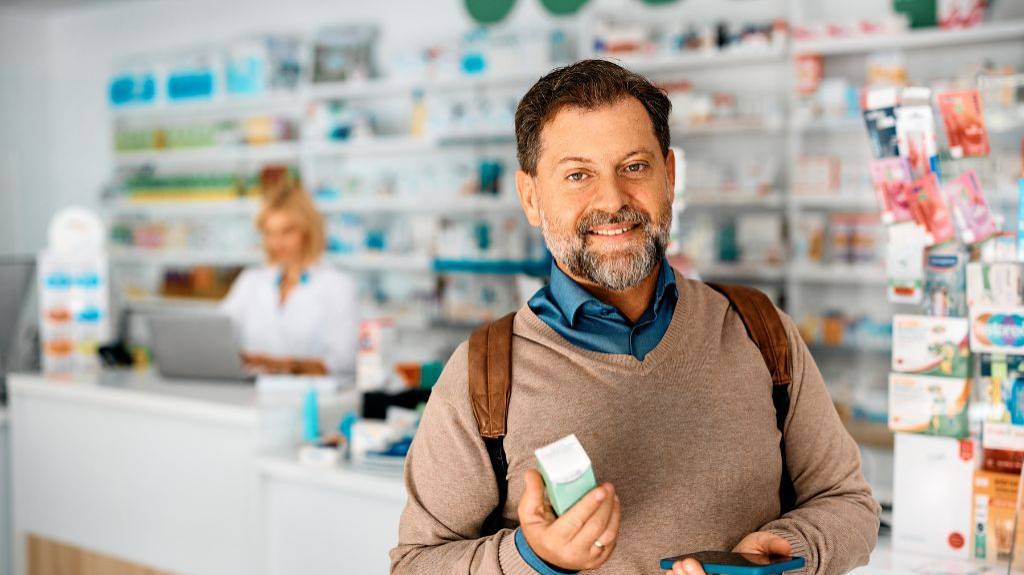 Ein Mann kauft ein Medikament in einer Apotheke. Er hält die Packung und sein Handy in der Hand. Im Hintergrund steht die Apothekerin am Tresen, außerdem viele Regale mit Medikamenten.