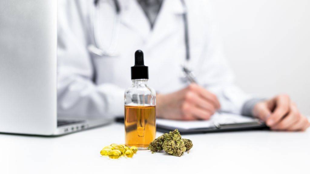 Kapseln, Cannabisblüten und Öl auf einem weißen Tisch. Im Hintergrund sitzt eine Person im Arztkittel neben einem Laptop und schreibt auf einem Klemmbrett.