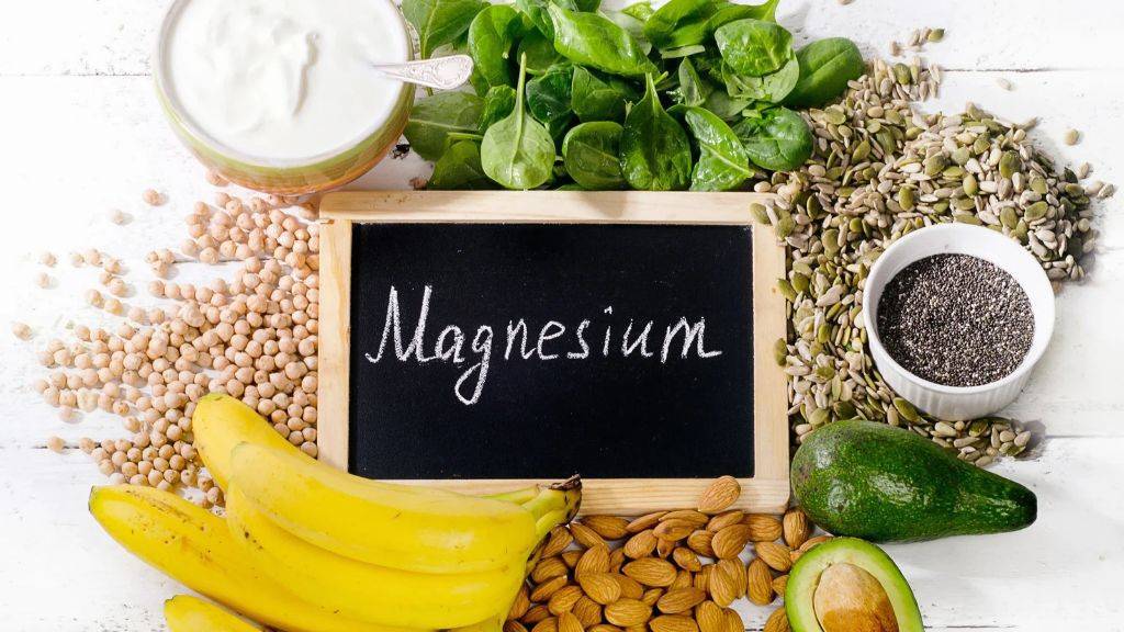 Lebensmittel, mit viel Magnesium auf einem weißen Holztisch. Joghurt, Banane, Spinat, Saaten, Nüsse und Avocado liegen um eine kleine schwarze Tafel mit Aufschrift: Magnesium.