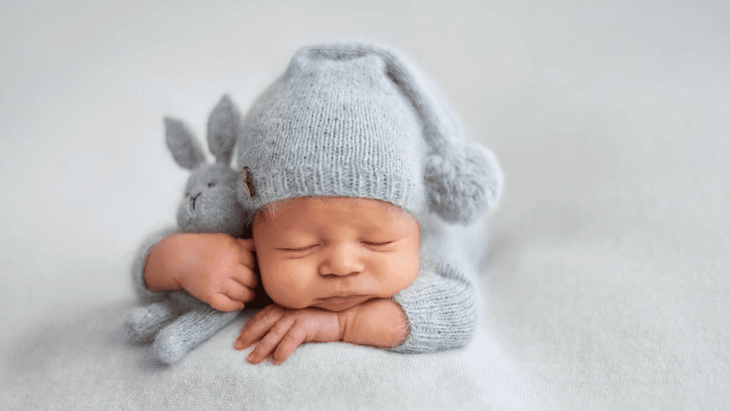 Neugeborenes schläft friedlich auf weicher Decke, trägt hellgraue Strickmütze mit Bommel und kuschelt mit passendem Stofftier – Symbol für Geborgenheit, Ruhe und die ersten Wochen mit Baby.