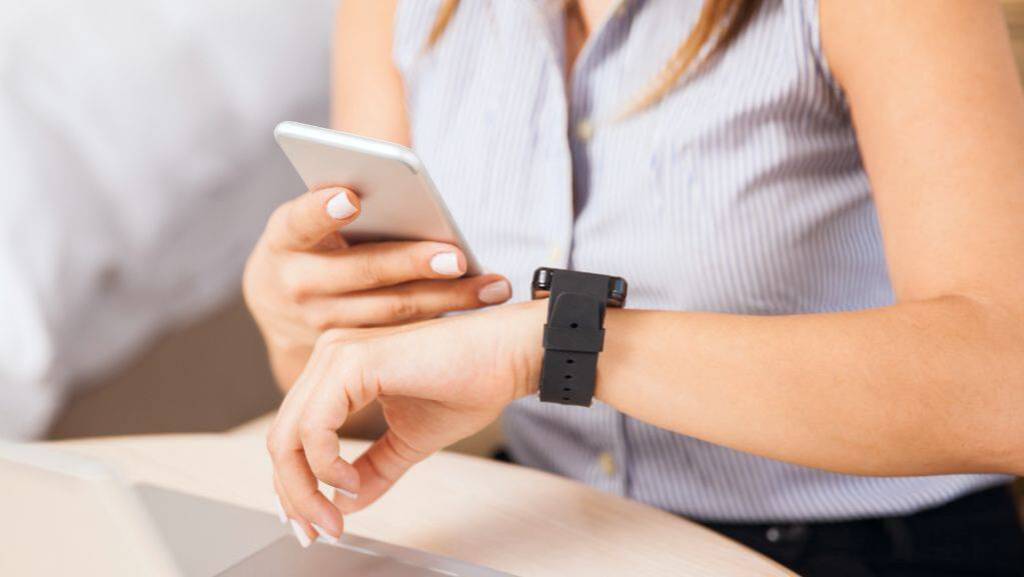 Ein Frau sitzt am Schreibtisch vor einem Laptop. Sie blickt auf Ihr Smartphone und ihre Smartwatch.
