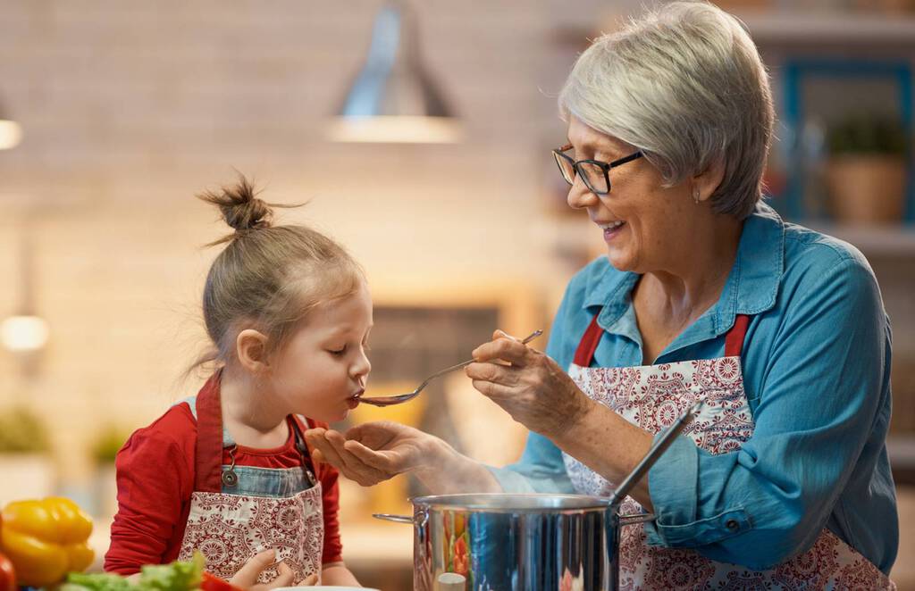Kind probiert Essen, dass Oma mit einen großen Löffel vor den Mund hält. Vor ihnen steht ein dampfender Topf.