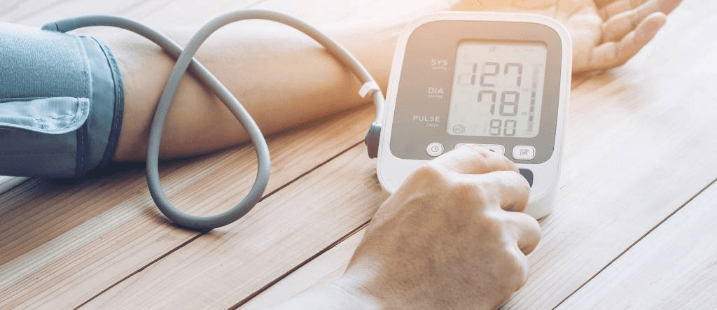 Person misst den Blutdruck mit Oberarmmanschette und digitalem Blutdruckmessgerät auf Holztisch, Anzeige mit systolischem und diastolischem Wert sowie Puls.