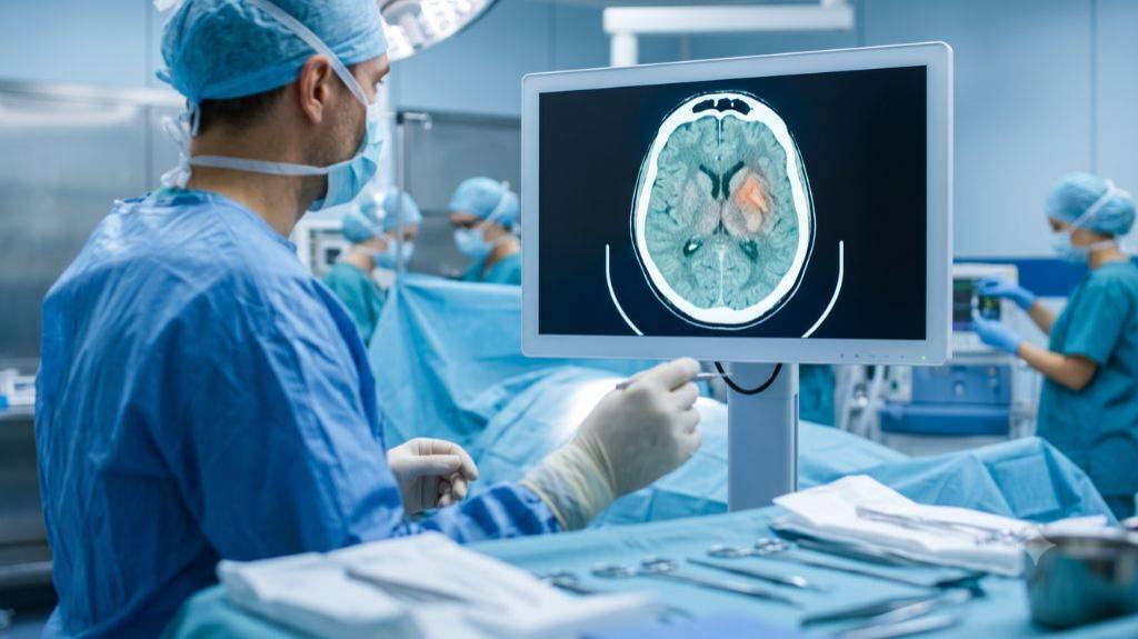 Blick über die Schulter eines Chirurgen in OP-Kleidung auf einen großen Monitor im OP-Saal. Der Bildschirm zeigt eine detaillierte CT-Aufnahme eines menschlichen Gehirns mit einer rot hervorgehobenen Läsion. Im Vordergrund liegen chirurgische Instrumente bereit, im Hintergrund ist weiteres medizinisches Personal in blauer OP-Kleidung zu sehen.