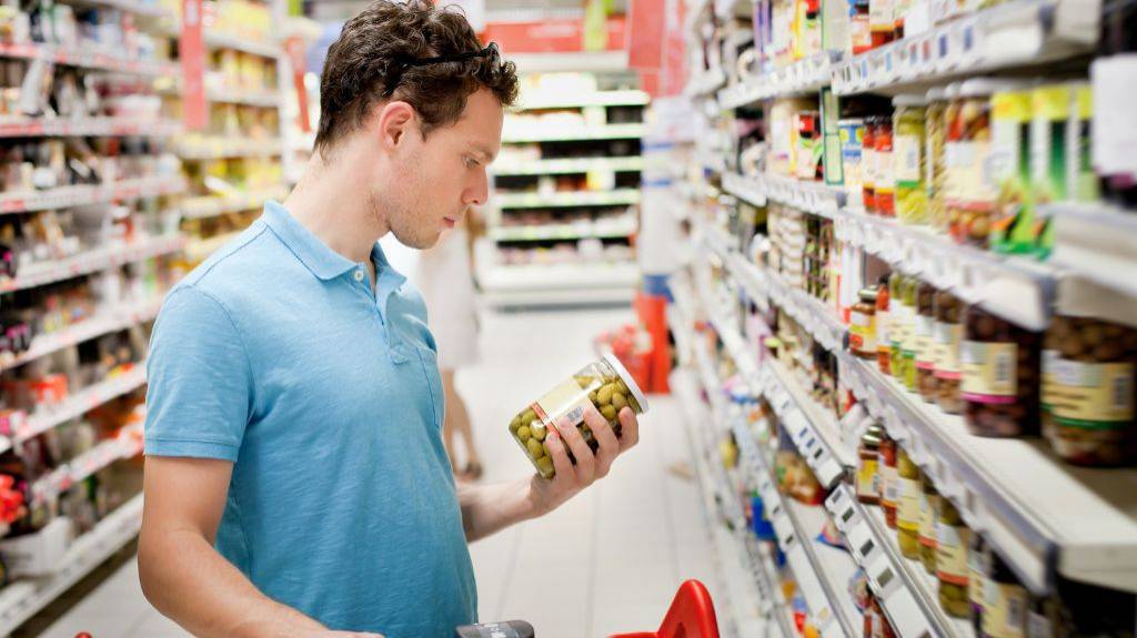 Ein Mann liest die Zutatenliste eines Glas eingelegter Oliven vor dem Supermarkt-Regal.