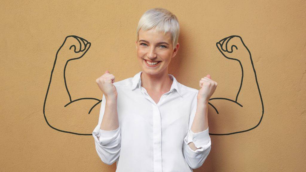 Sportliche ältere Frau mit kurzen grauen Haaren in motivierender Pose, Hände zu Fäusten geballt, Arme gebeugt und nah am Körper. Im Hintergrund an einer bräunlichen Wand eine Zeichnung von zwei Armen, die den Bizeps zeigen.n