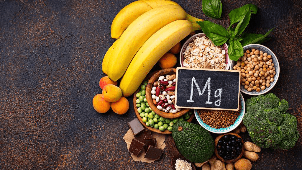 Magnesiumreiche Lebensmittel wie Bananen, Haferflocken, Hülsenfrüchte, Brokkoli, Avocado, Nüsse und Schokolade mit Mg-Tafel auf dunklem Hintergrund.