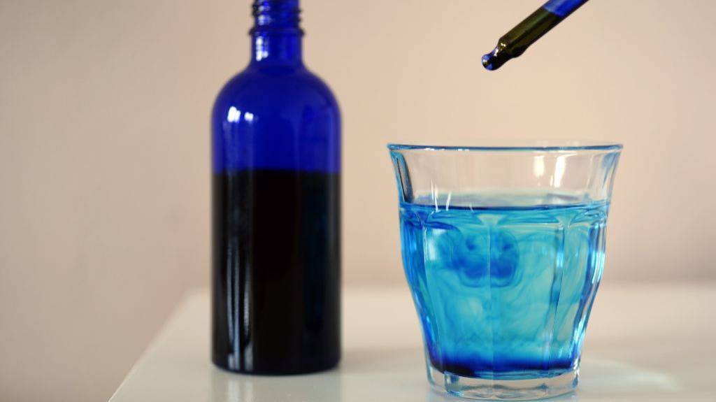 Eine Pipette tropft Methylenblau in ein kleines Wasserglas. Im Hintergrund steht eine blaue Flasche. 