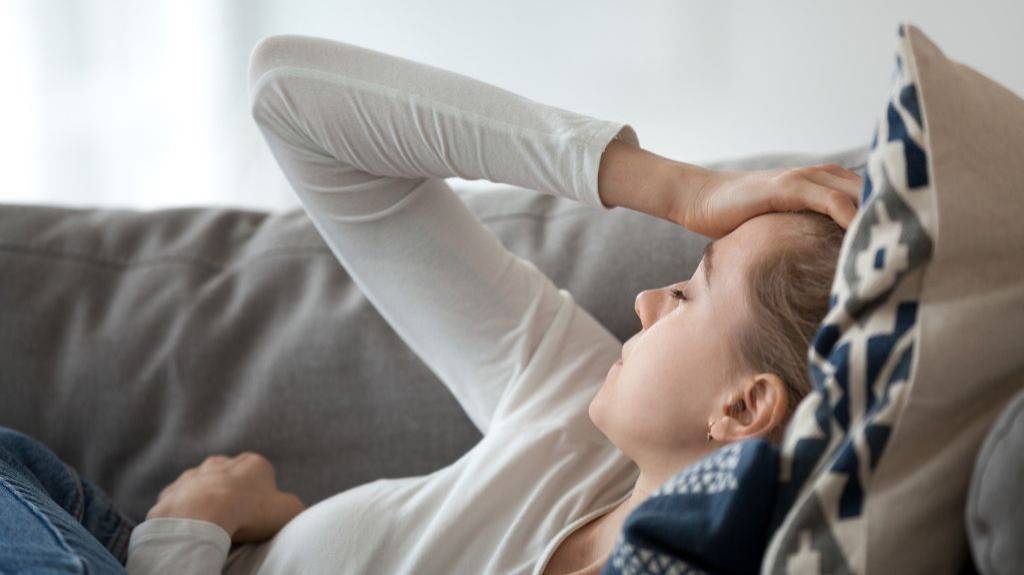 Frau liegt mit geschlossenen Augen auf der Couch und hält sich den Kopf.
