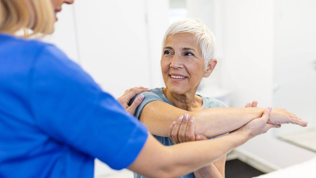 Frau mit grauen kurzen Haaren sitzt auf einer Liege und führt mit Unterstützung einer Physiotherapeutin eine Dehnübung des Armes durch.