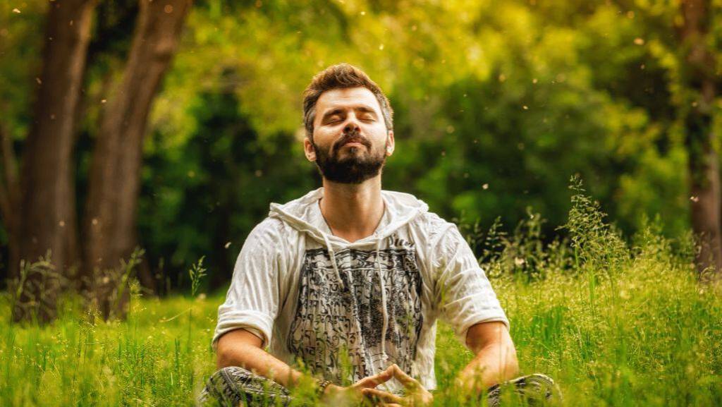 Ein Mann sitzt auf einer Waldlichtung im hohen Gras und meditiert.