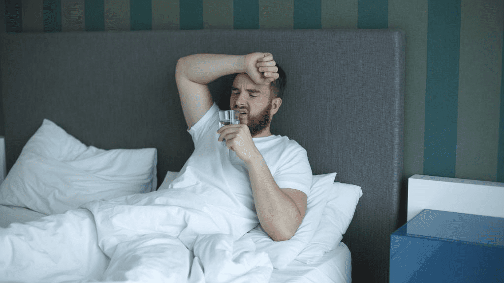 Ein Mann mit Bart liegt in einem Bett, hält ein Glas Wasser in der Hand und sieht erschöpft aus. 