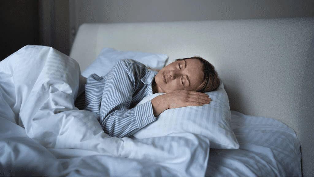 Frau schläft entspannt in einem Bett mit weißer Bettwäsche, trägt ein blau-weiß gestreiftes Schlafhemd und liegt auf der Seite mit dem Kopf auf einem Kissen – Symbol für gesunden und erholsamen Schlaf.