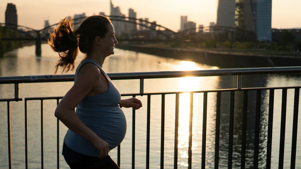 Eine schwangere Frau beim Joggen. Sie läuft über eine Brücke. Man sieht die tiefstehende Sonne, einen breiten Fluss und eine Stadt im Hintergrund.