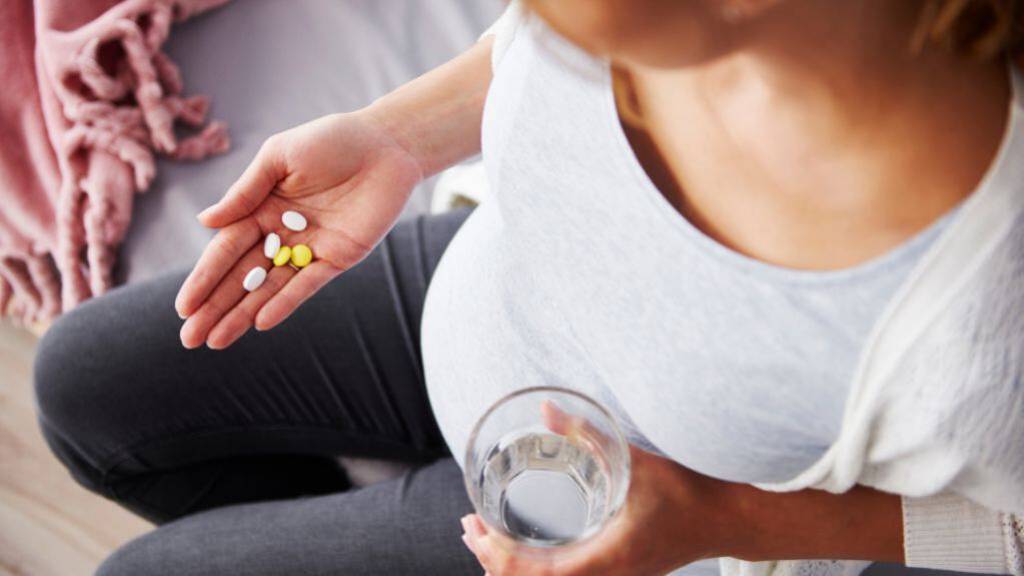 Eine Schwangere hält mehrere Tabletten in der Handfläche, in der anderen Hand hat sie ein Wasserglas. Ungewöhnliche Perspektive: Blick über die Schulter von oben.