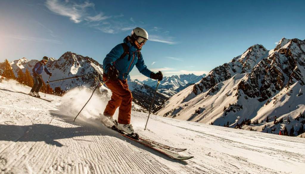 Ein Skifahrer in blauer Jacke und oranger Hose fährt dynamisch eine perfekt präparierte Piste hinunter. Im Hintergrund erheben sich majestätische, schneebedeckte Berge unter blauem Himmel und Sonnenschein.