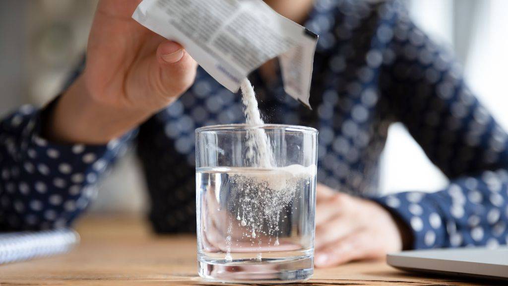 Eine Frau schüttet weißes Punlver aus einer Medikamentenpackung in ein Wasserglas.