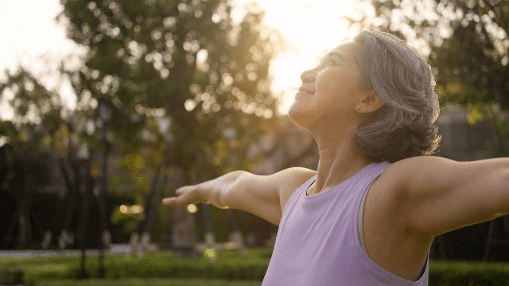 Frau mittleren Alters steht entspannt im Park, Arme ausgebreitet und Gesicht zur Sonne. Symbolbild für Wohlbefinden, neue Lebensphase und ganzheitliche Begleitung in den Wechseljahren.